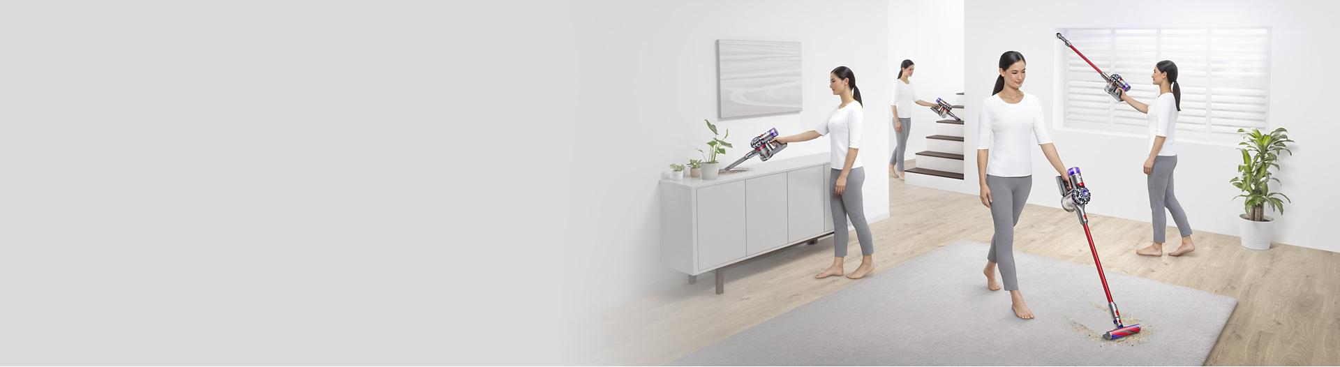 Wide shot of a woman cleaning all over her living room with a Dyson V8 vacuum as a dog walks in.