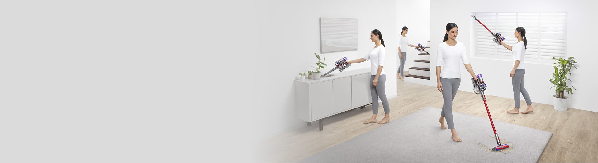 Wide shot of a woman cleaning all over her living room with a Dyson V8 vacuum as a dog walks in.