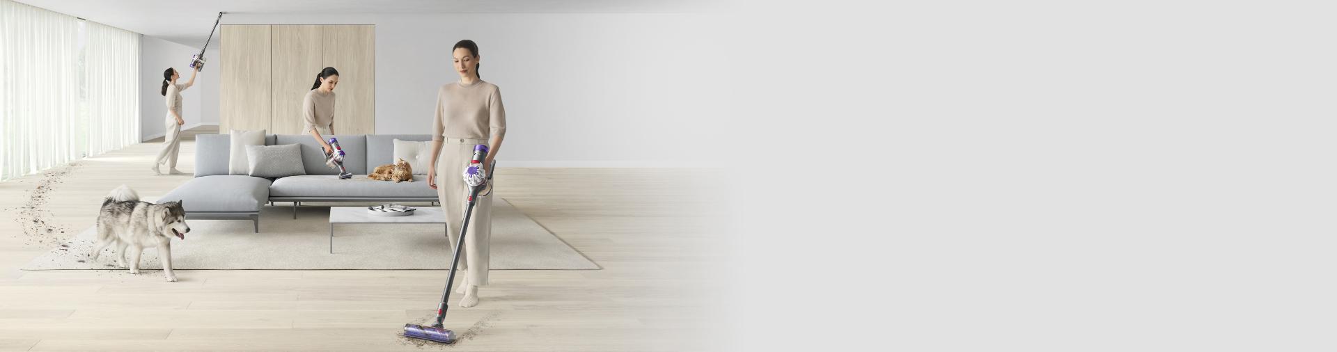 لقطة عريضة لسيدة تُنظّف غرفة الجلوس بالكامل باستخدام مكنسة Dyson V8 بينما يدخل كلبها.