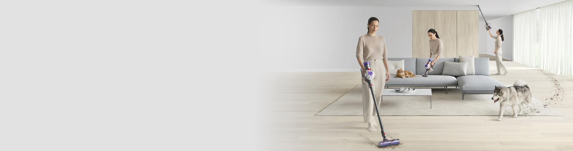 Wide shot of a woman cleaning all over her living room with a Dyson V8 vacuum as a dog walks in.