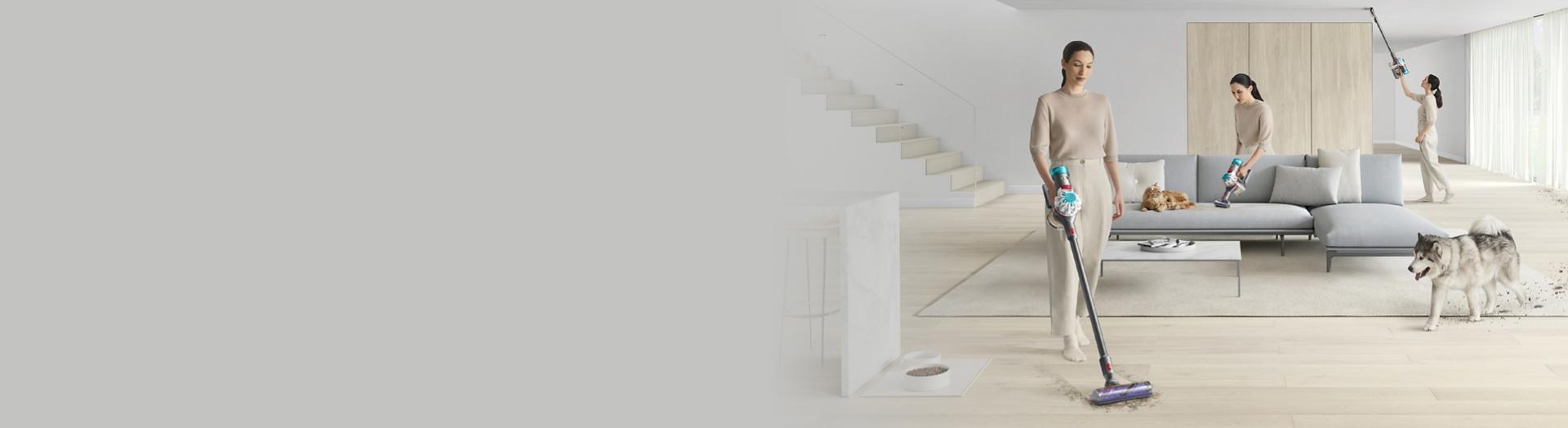 Wide shot of a woman cleaning all over her living room with a Dyson V8 vacuum as a dog walks in.