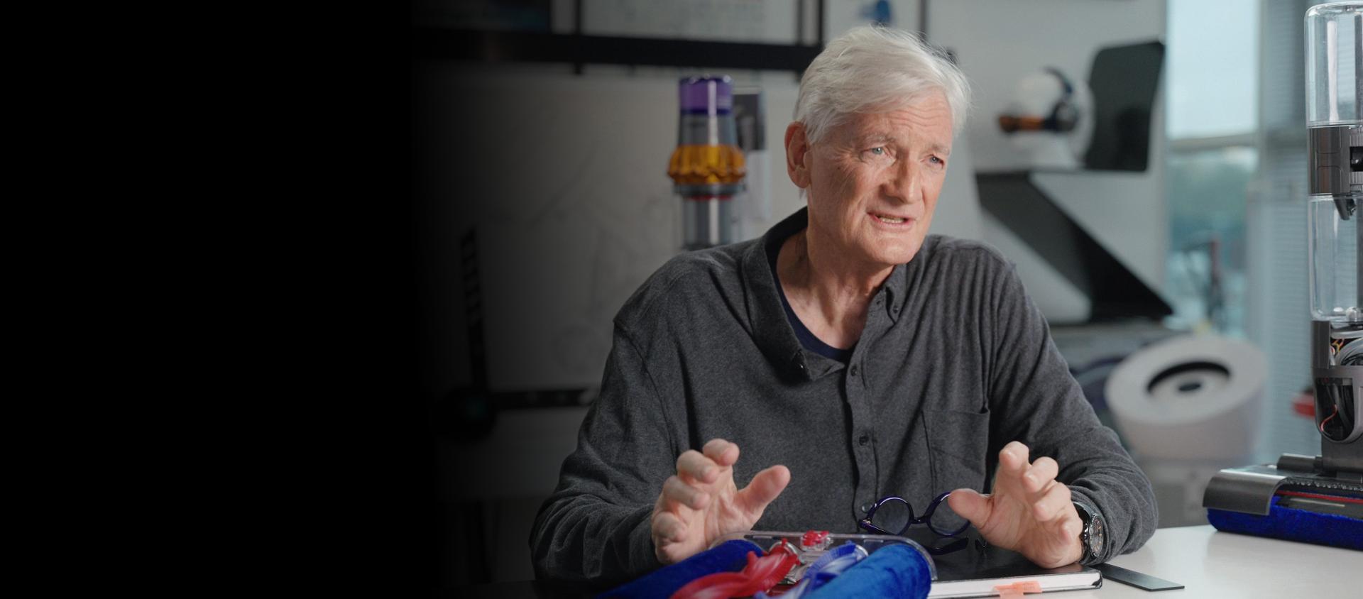James Dyson siedzi w swoim biurze, otoczony produktami i częściami Dyson.
