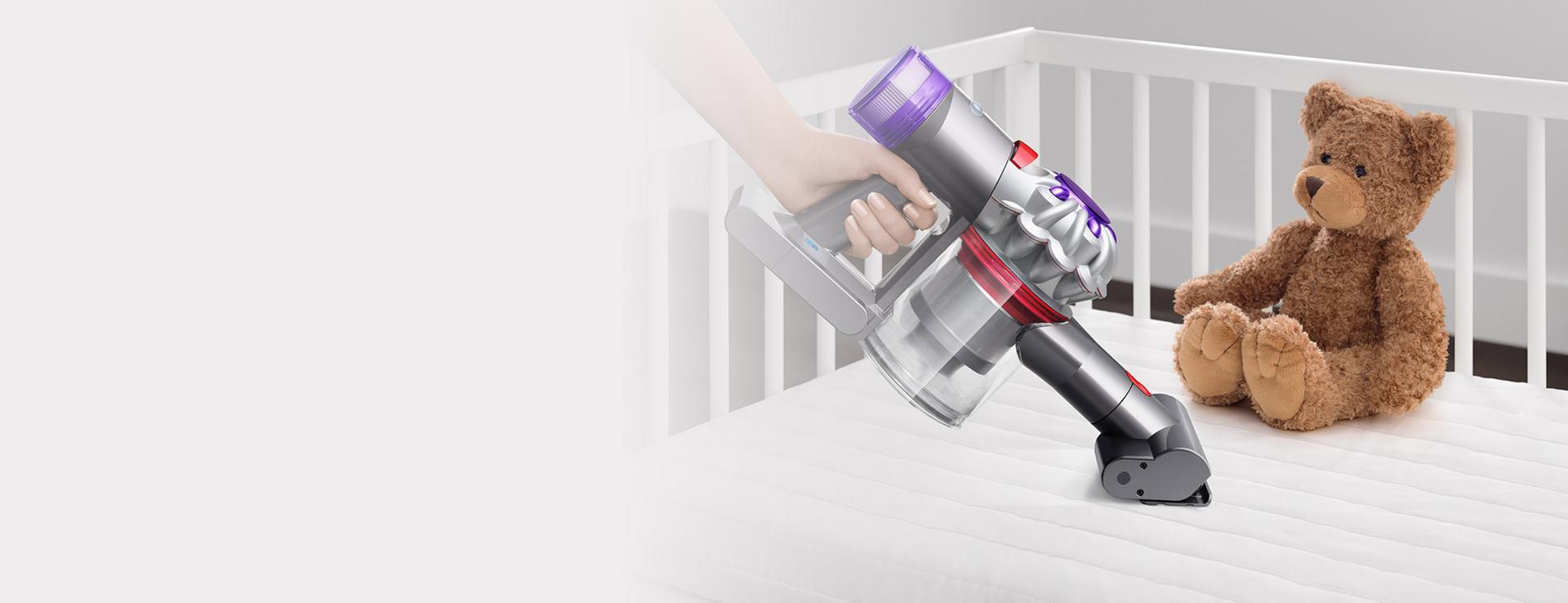 A person uses the Mini motorised head on the Car+Boat vacuum to clean a baby's cot mattress with a teddy bear in the background.