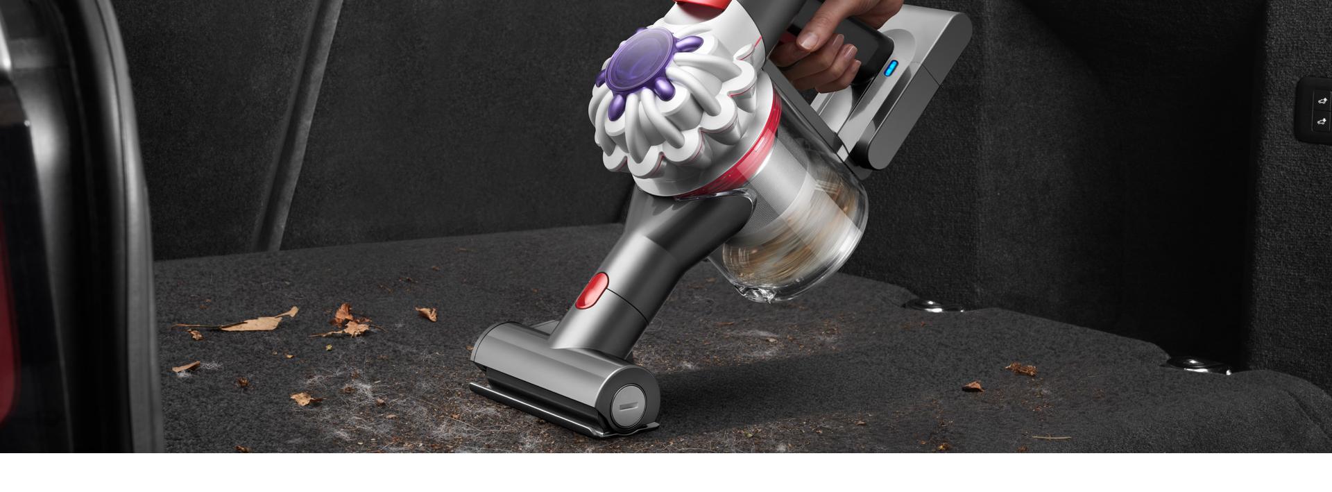 A Dyson Car+Boat handheld vacuum cleaner with the Mini motorised tool is used to clean pet hair and other debris from a car boot carpet.