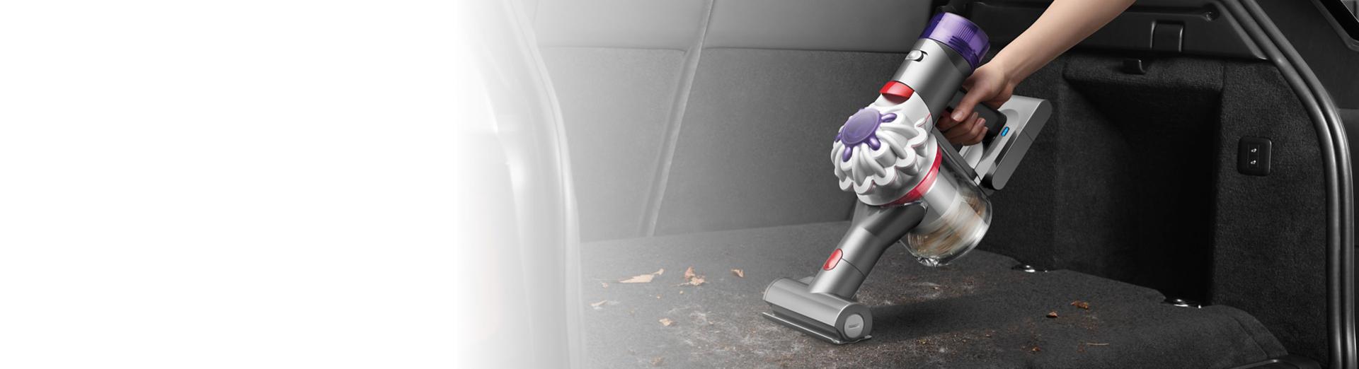 A Dyson Car+Boat handheld vacuum cleaner with the Mini motorised tool is used to clean pet hair and other debris from a car boot carpet.