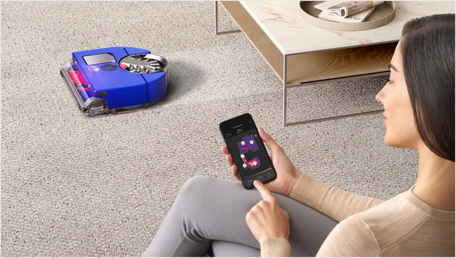 Woman on a sofa using the MyDyson™ app, with the robot in the background