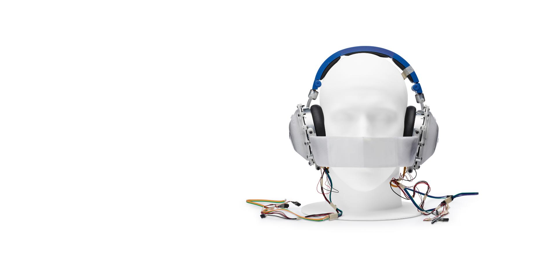 Manikin head wearing prototype headphones, with band across nose and mouth, and wires coming from the earcups