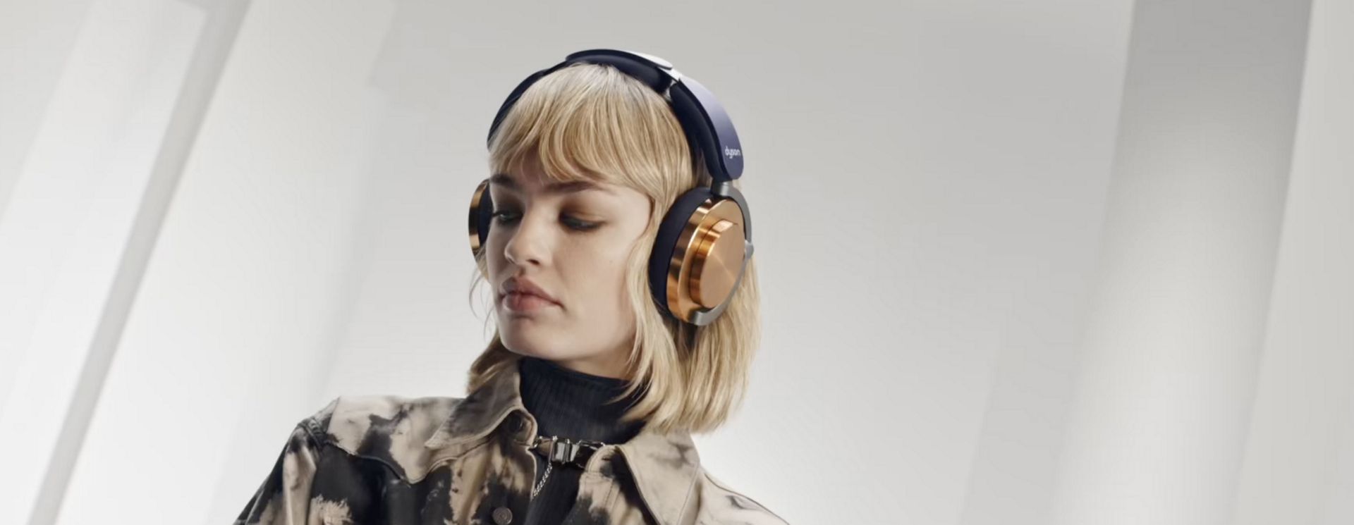 Šviesiaplaukė moteris modelis, žiūrinti žemyn ir dėvinti ausines „Dyson OnTrac“ su CNC vario kaušeliais.