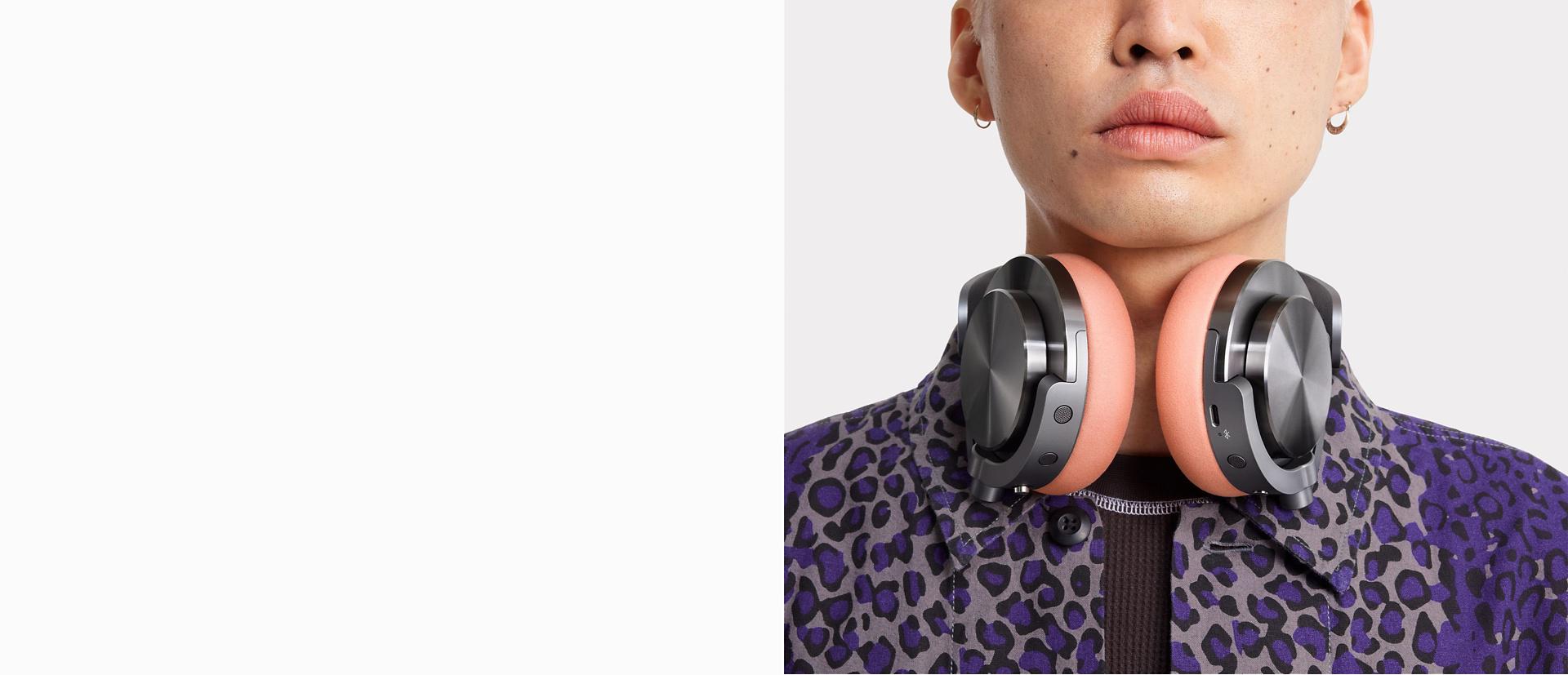 CNC 블랙 니켈 캡과 오이스터 핑크 이어쿠션을 장착한 다이슨 온트랙 헤드폰을 목에 걸고 카메라를 바라보는 모델 근접 이미지.