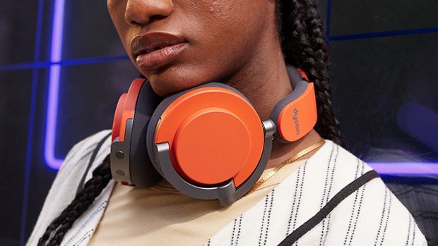 Modelis, ant kaklo užsidėjęs ausines „Dyson OnTrac“ keramikos cinoberio spalvos išoriniais ausų kaušeliais.