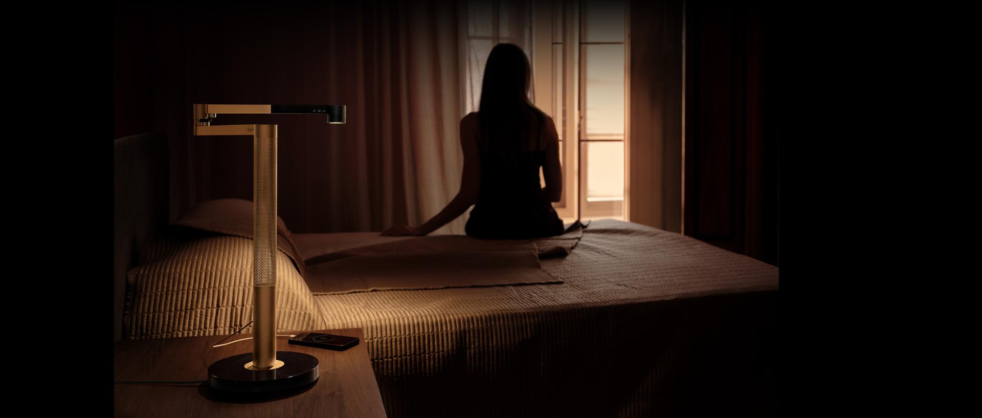 komodinin üzerinde Dyson Solarcycle Morph masa lambası ile yatağının kenarında oturan ürün sahibi.
