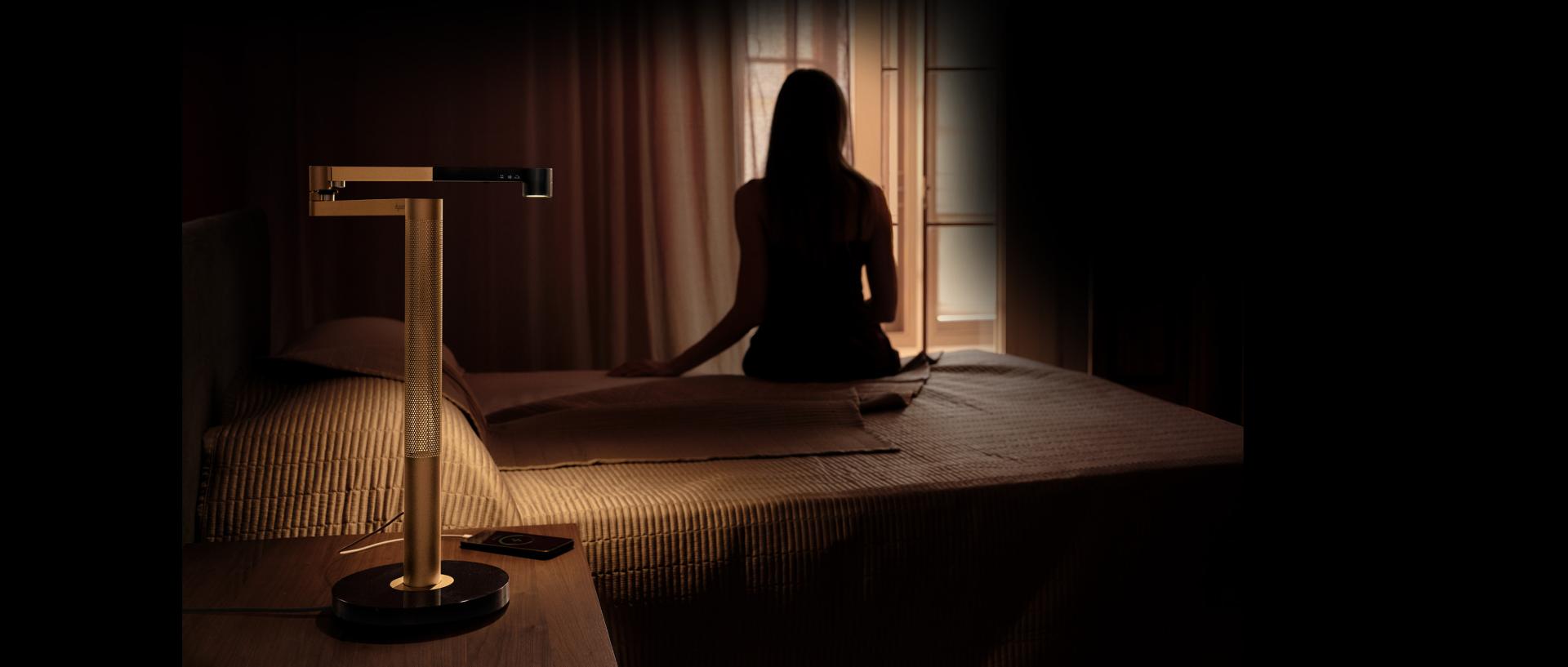 Propietaria sentada en el borde de la cama con la lámpara de escritorio Dyson Solarcycle Morph en la mesilla de noche.