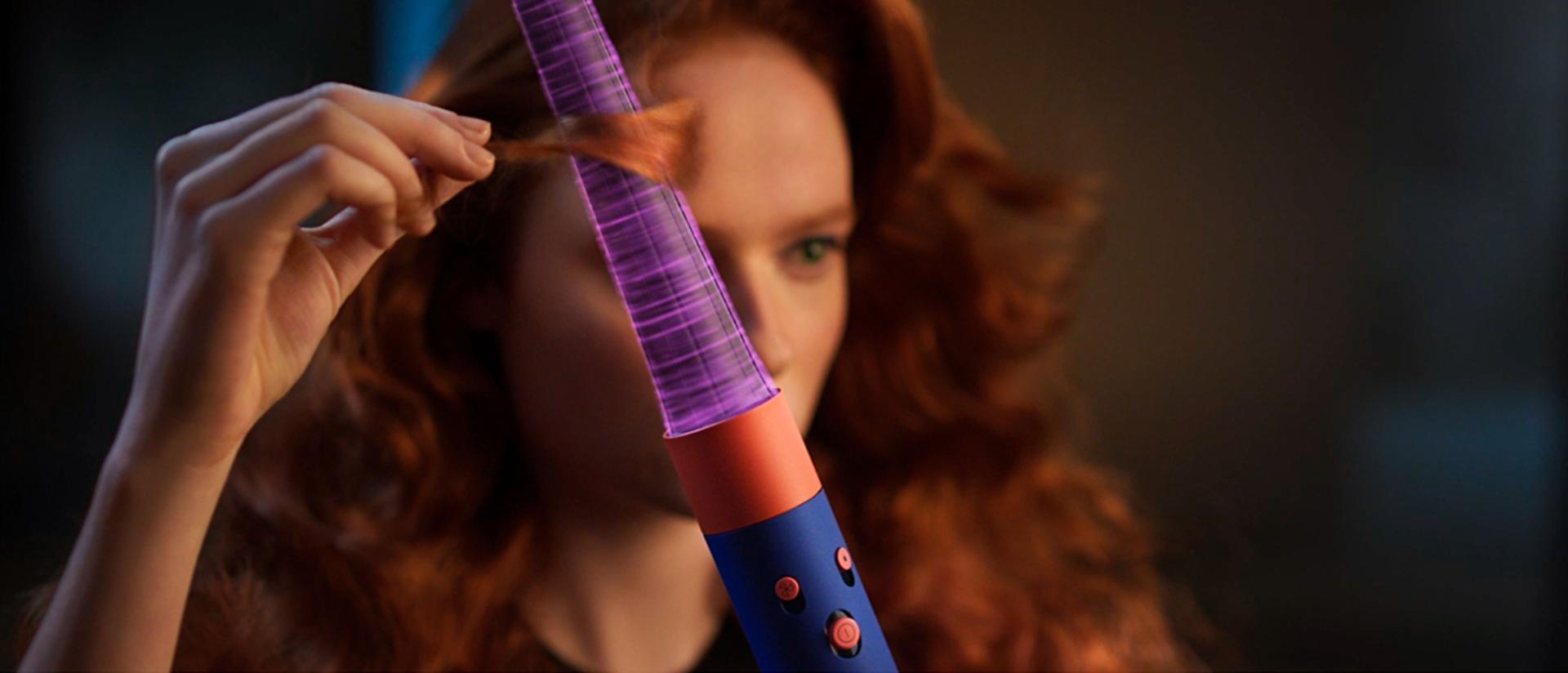 A redhead model wraps a tress of her hair around the Conical Airwrap barrel with pink graphics showing the Coanda airflow.