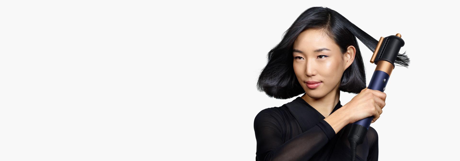 Un model cu păr negru de lungime medie folosește accesoriul de netezire pentru firele rebele și uscător pentru a-și netezi coafura.