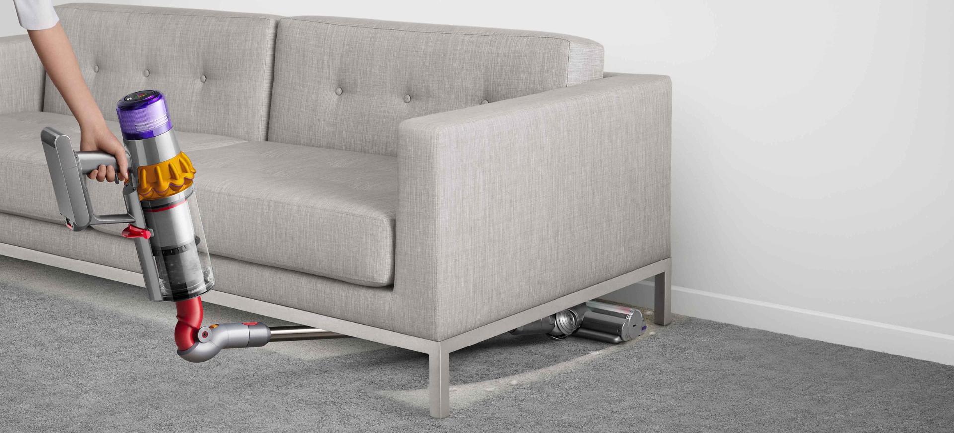 مكنسة Dyson V15 Detect في مهمة تنظيف تحت أريكة رمادية.