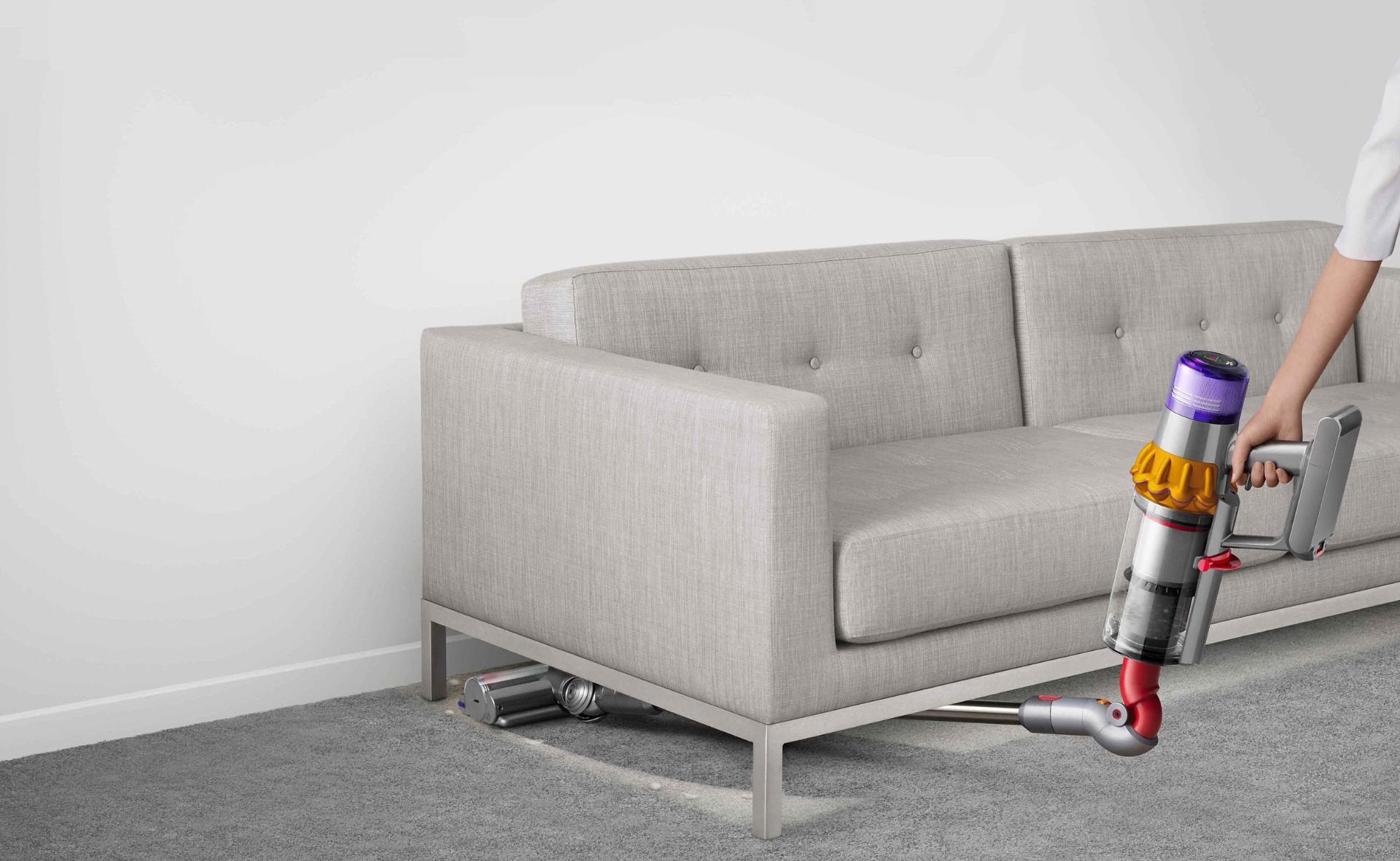 Dyson V15 Detect vacuum vacuuming under a grey sofa.