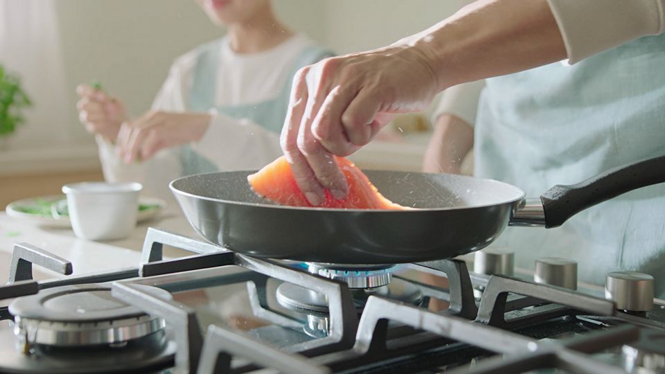 Eine Hand bereitet Lachs in einer Pfanne auf einem Gasherd zu.