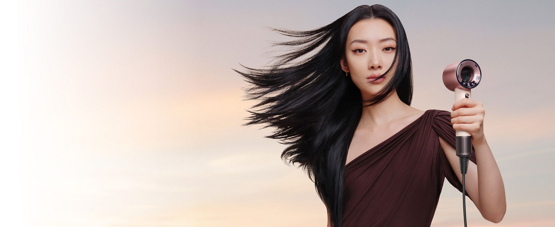 A female model with black straight and wavy hair holds the Dyson Supersonic Travel hair dryer.