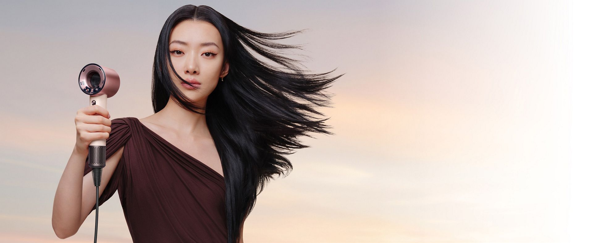 A female model with black straight and wavy hair holds the Dyson Supersonic Travel hair dryer.