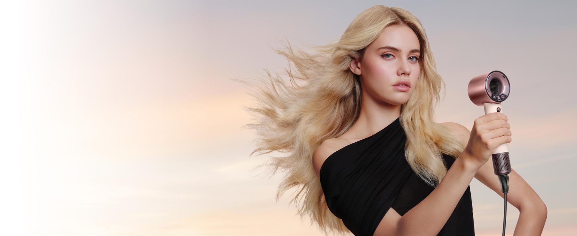 A female model with blonde straight and wavy hair holds the Dyson Supersonic Travel hair dryer.