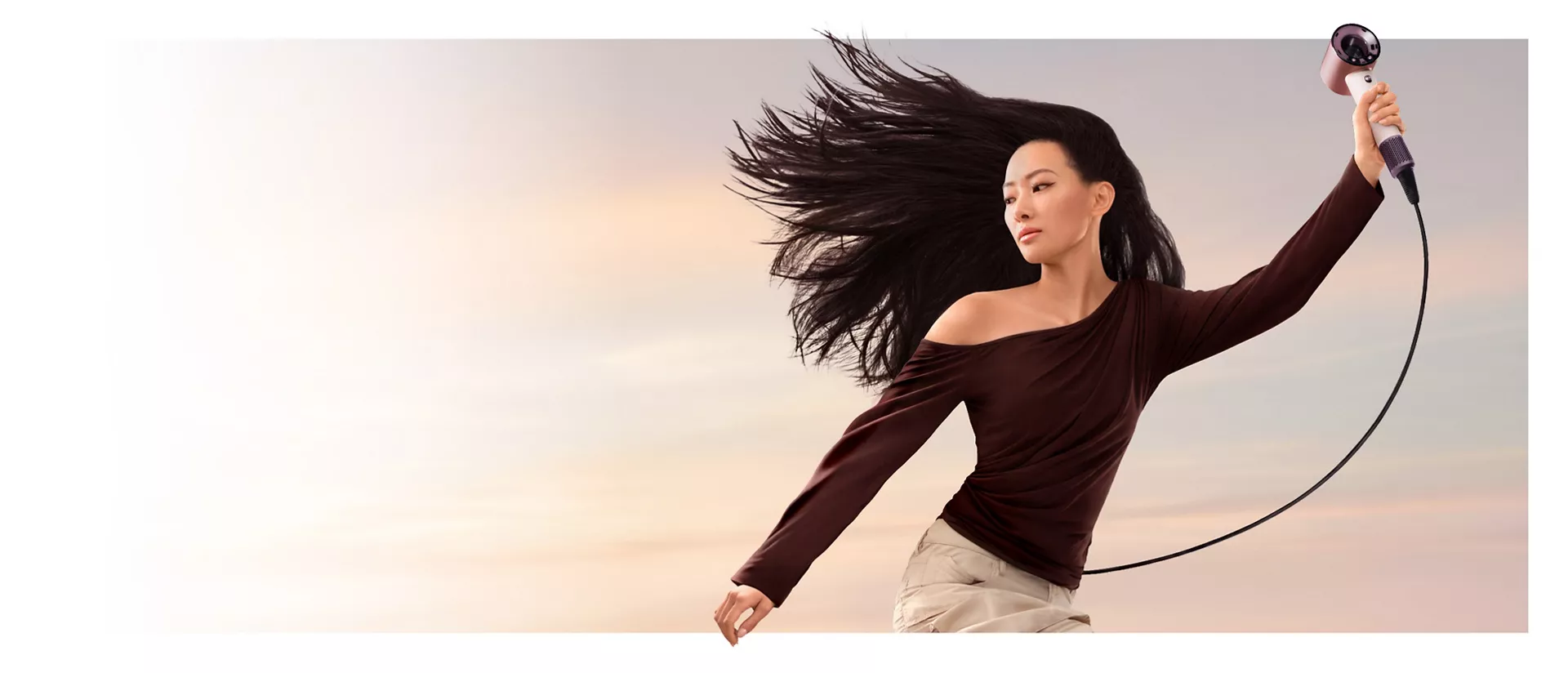 A jumping female model in mid-air holding the Dyson Supersonic Travel hair dryer.