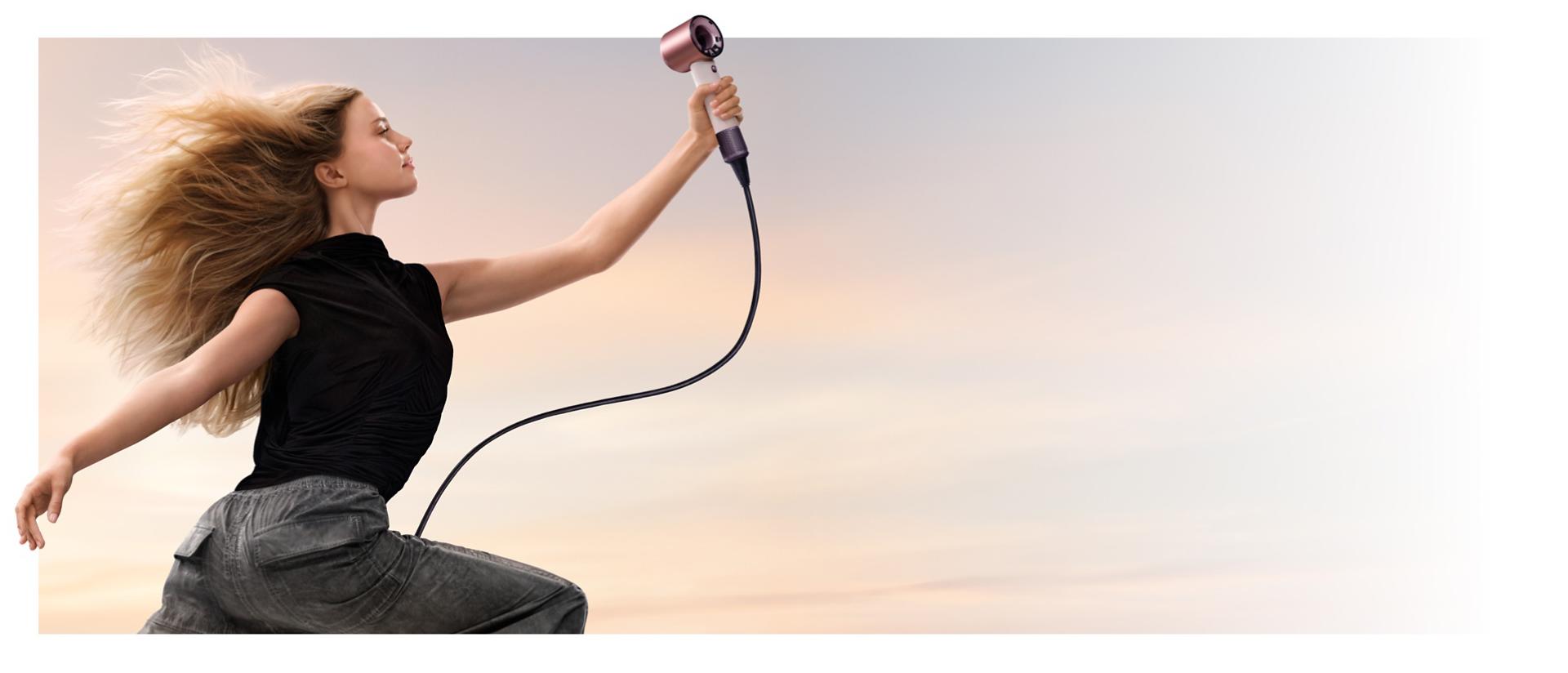 A jumping female model in mid-air holding the Dyson Supersonic Travel hair dryer.