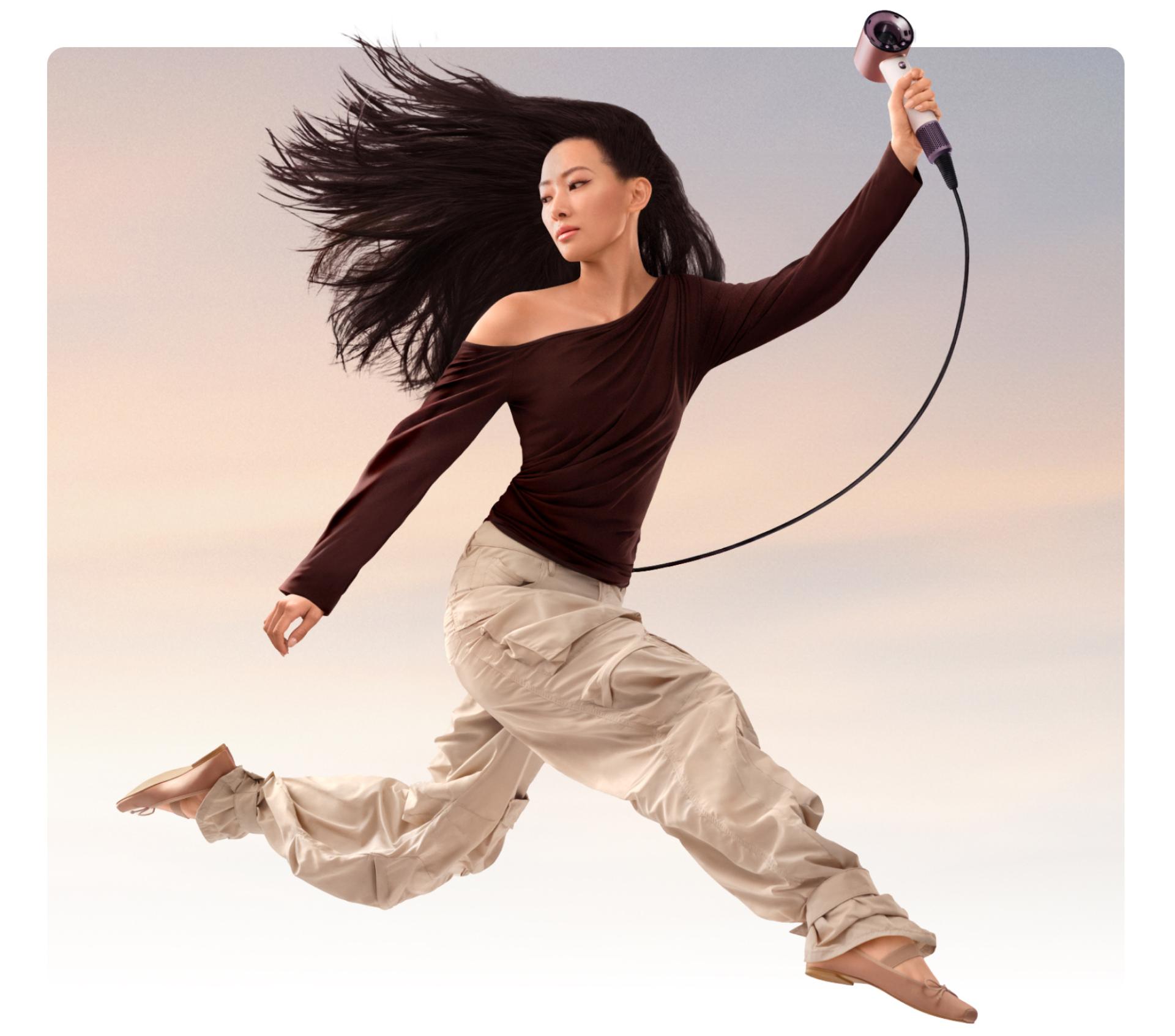 A jumping female model in mid-air holding the Dyson Supersonic Travel hair dryer.