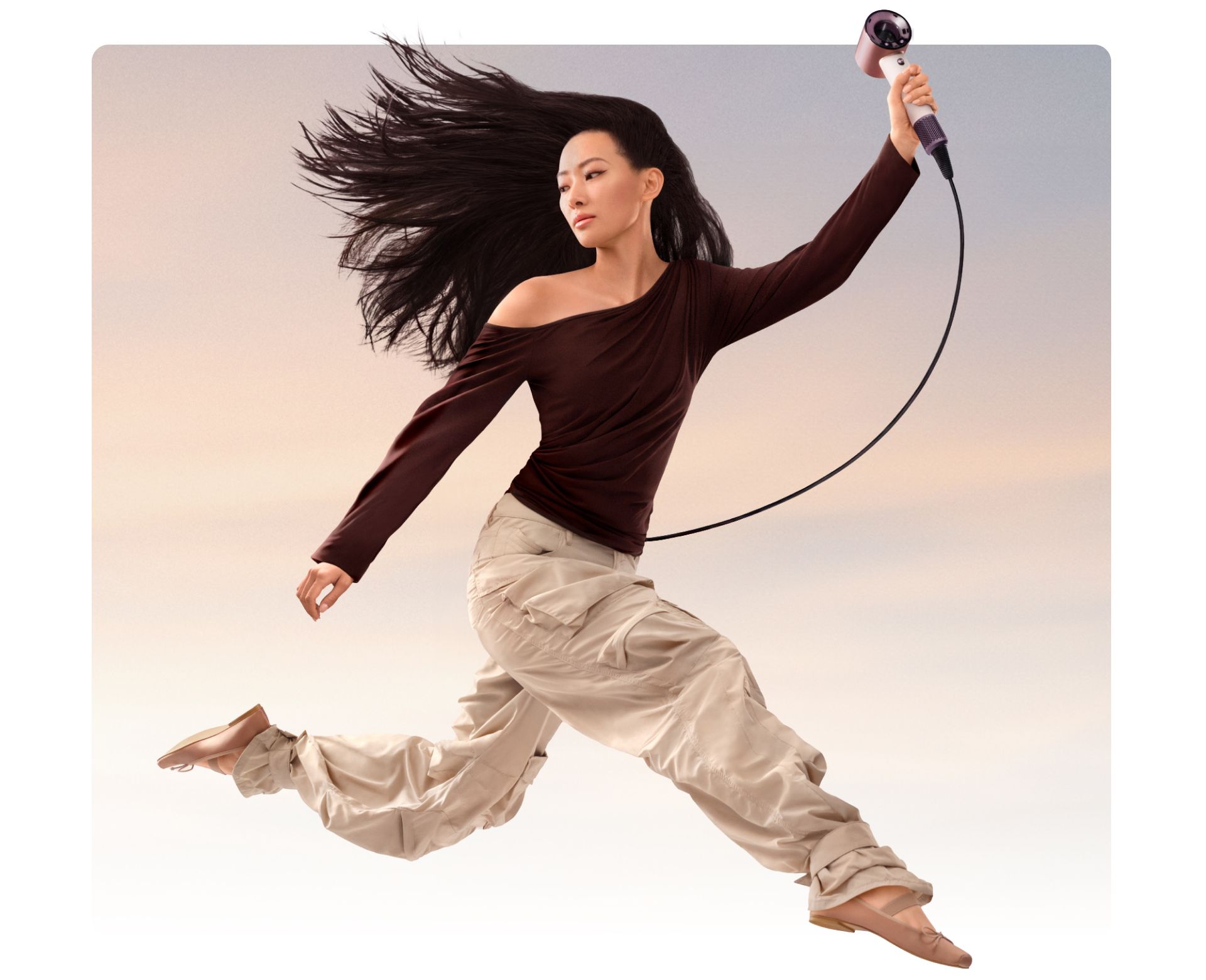 A jumping female model in mid-air holding the Dyson Supersonic Travel hair dryer.
