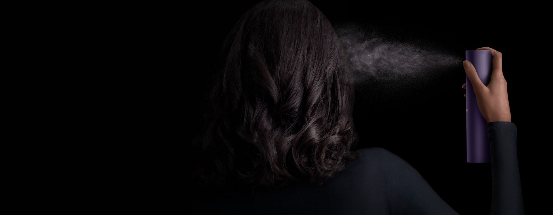 짙은 머리카락을 가진 모델이 다이슨 키토산 멀티유즈 스타일링 스프레이를 자신의 머리에 바릅니다.