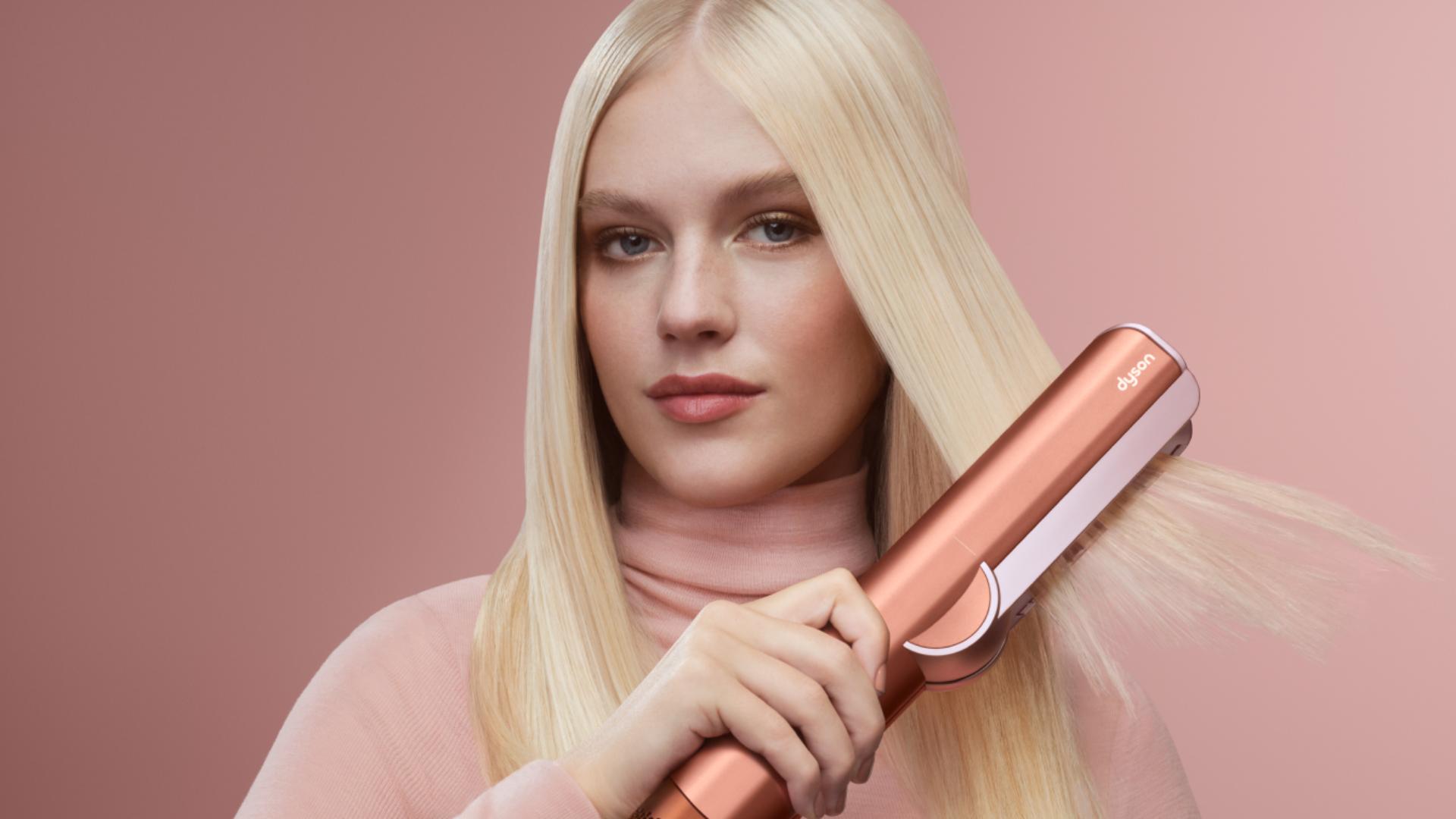 Blond modelka si vysouší a upravuje vlasy pomocí vysoušeče a žehličky na vlasy Dyson Airstrait.