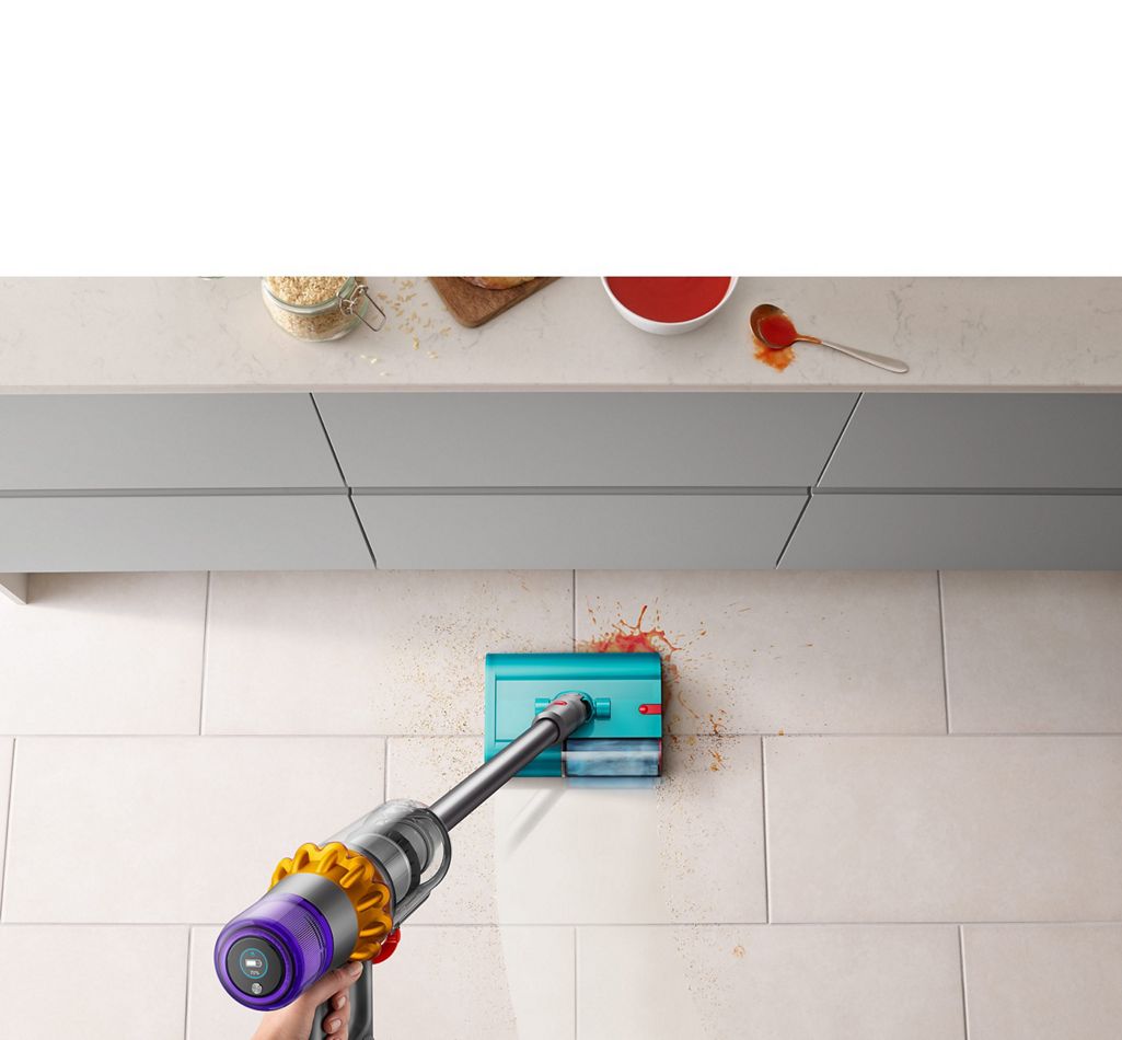 Dyson V15s Detect Submarine wet roller head clearing up spilt soup and breadcrumbs on a tiled kitchen floor near a countertop.
