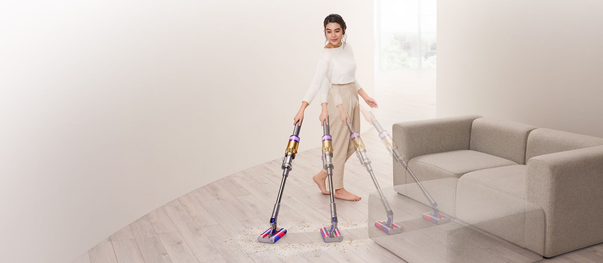 Frau reinigt mit dem Dyson Omni-glide™ Staubsauger auf einem Hartboden um ein Sofa herum.