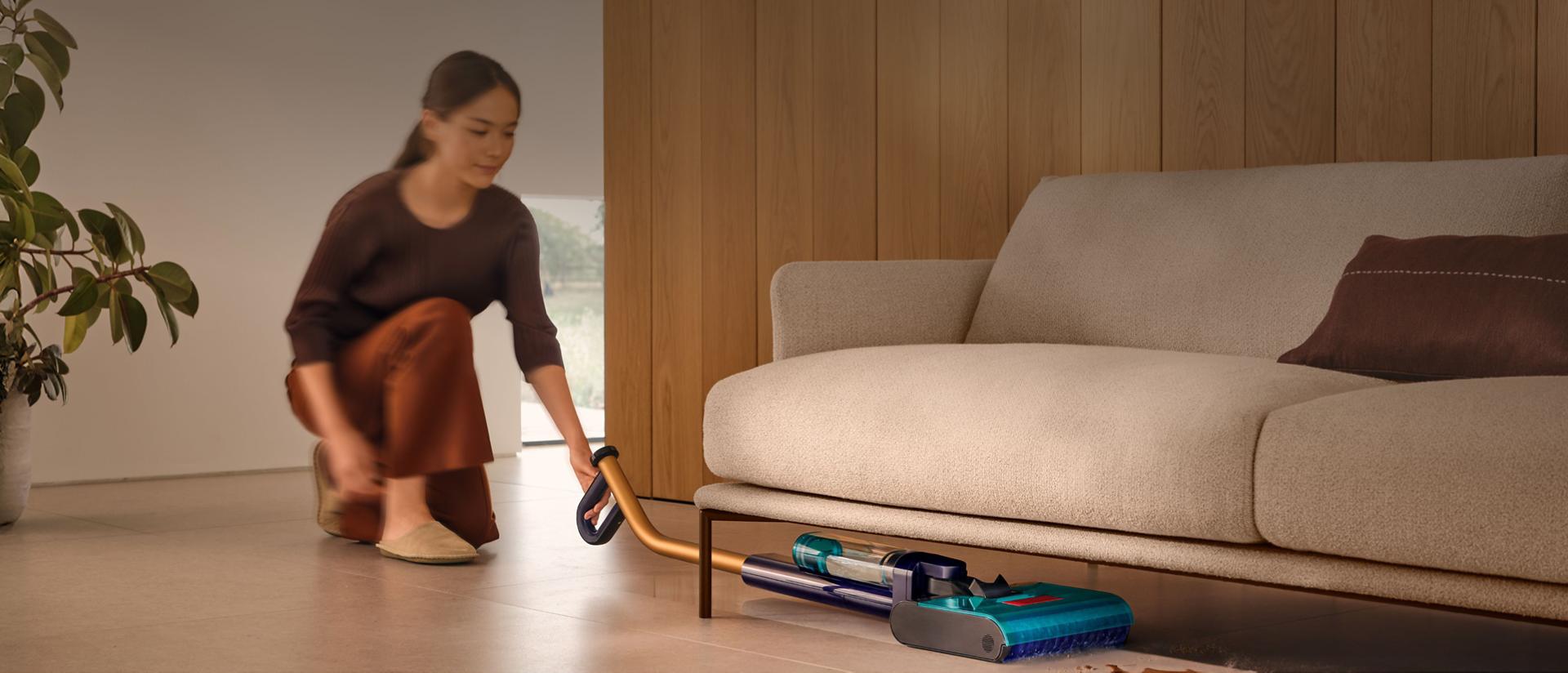 يُنظِّف جهاز تنظيف الأرضيات الصلبة Dyson Clean+Wash Hygiene تحت أريكة.