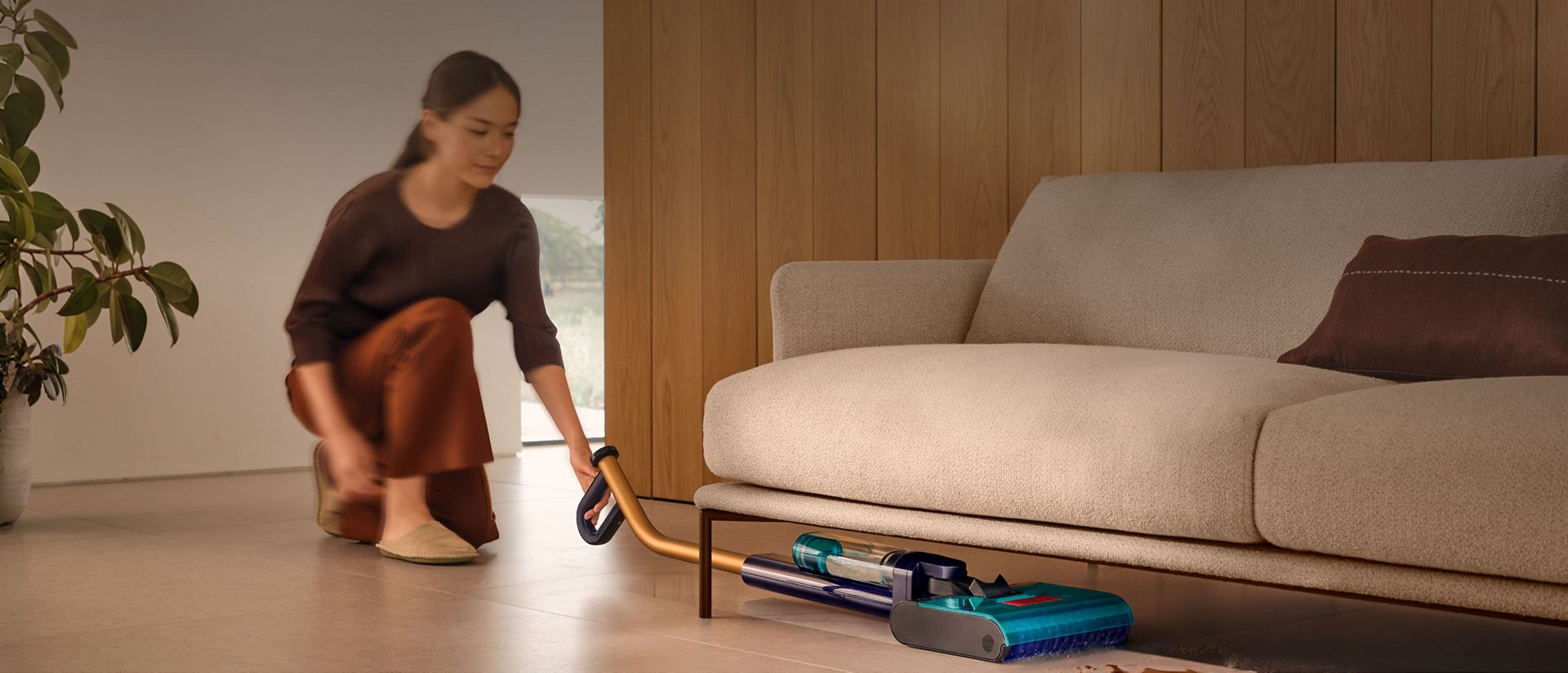 يُنظِّف جهاز تنظيف الأرضيات الصلبة Dyson Clean+Wash Hygiene تحت أريكة.