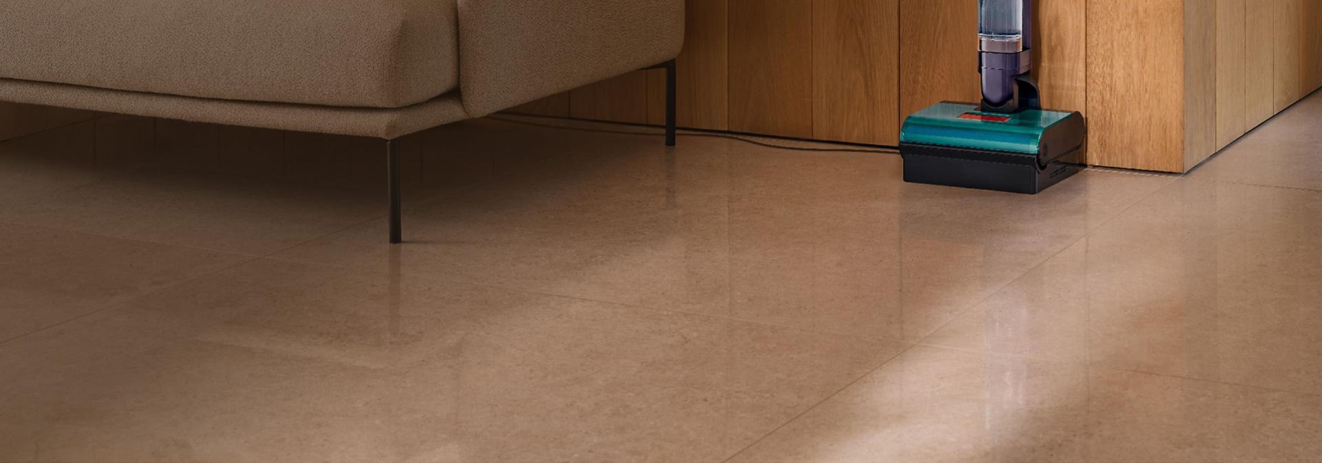 Wysokoobrotowy mop elektryczny Dyson Clean+Wash Hygiene w trakcie czyszczenia podłogi pod kanapą.