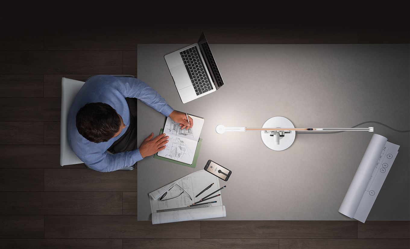  A top down view of a man working in a notebook on his desk, while the Dyson Solarcycle Morph desk lamp illuminates his work station.
