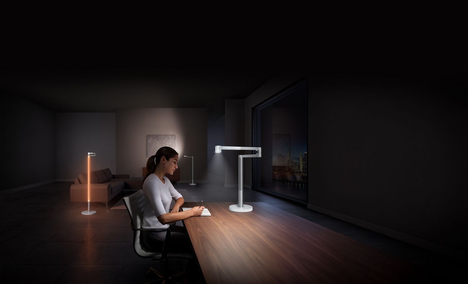 Une femme travaille proche d'une lampe de travail Dyson Solarcycle🅪 s’ajustant à la lumière de la nuit à l’extérieur d’une fenêtre