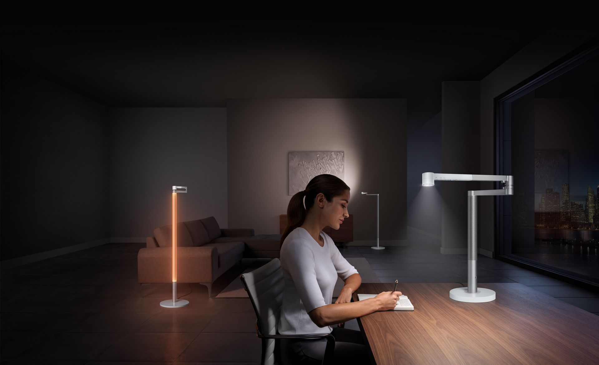 Une femme travaille proche d'une lampe de travail Dyson Solarcycle🅪 s’ajustant à la lumière de la nuit à l’extérieur d’une fenêtre